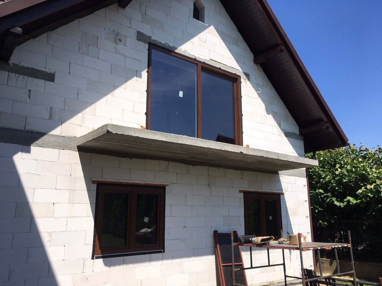 Surowy budynek z białego bloczka z zamontowanymi brązowymi oknami, widoczne rusztowanie i narzędzia na zewnątrz, etap budowy.