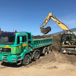 Żółta koparka CAT M315C nabiera ziemię do zielonej ciężarówki MAN z pomarańczowymi elementami na tle błękitnego nieba i szarego budynku.