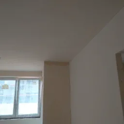 Świeżo położone, gładkie tynki maszynowe na ścianach i suficie w jasnym pomieszczeniu z oknem i widocznym narożnikiem ściany.