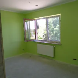 Pomalowane na jasnozielono wnętrze pokoju z otwartym oknem, grzejnikiem i wylewką na podłodze.