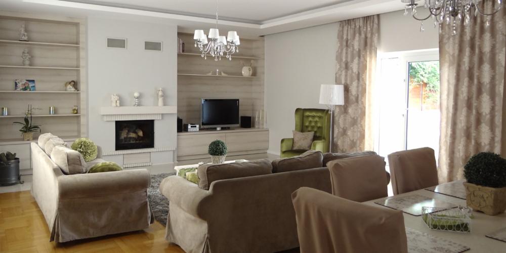 Jasne wnętrze salonu z kominkiem, beżową sofą, zielonym fotelem i kryształowym żyrandolem, widok na ogród przez okno balkonowe.