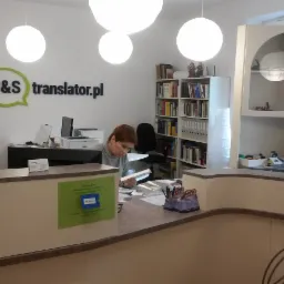 Biuro firmy tłumaczeniowej B&S translator.pl z pracującą kobietą nad dokumentami przy ladzie recepcyjnej, widoczny regał z książkami i drugi pracownik przy oknie.