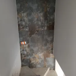 Ściana wyłożona płytkami imitującymi rdzę, widok z dołu schodów w trakcie prac, zabezpieczone taśmą malarską.