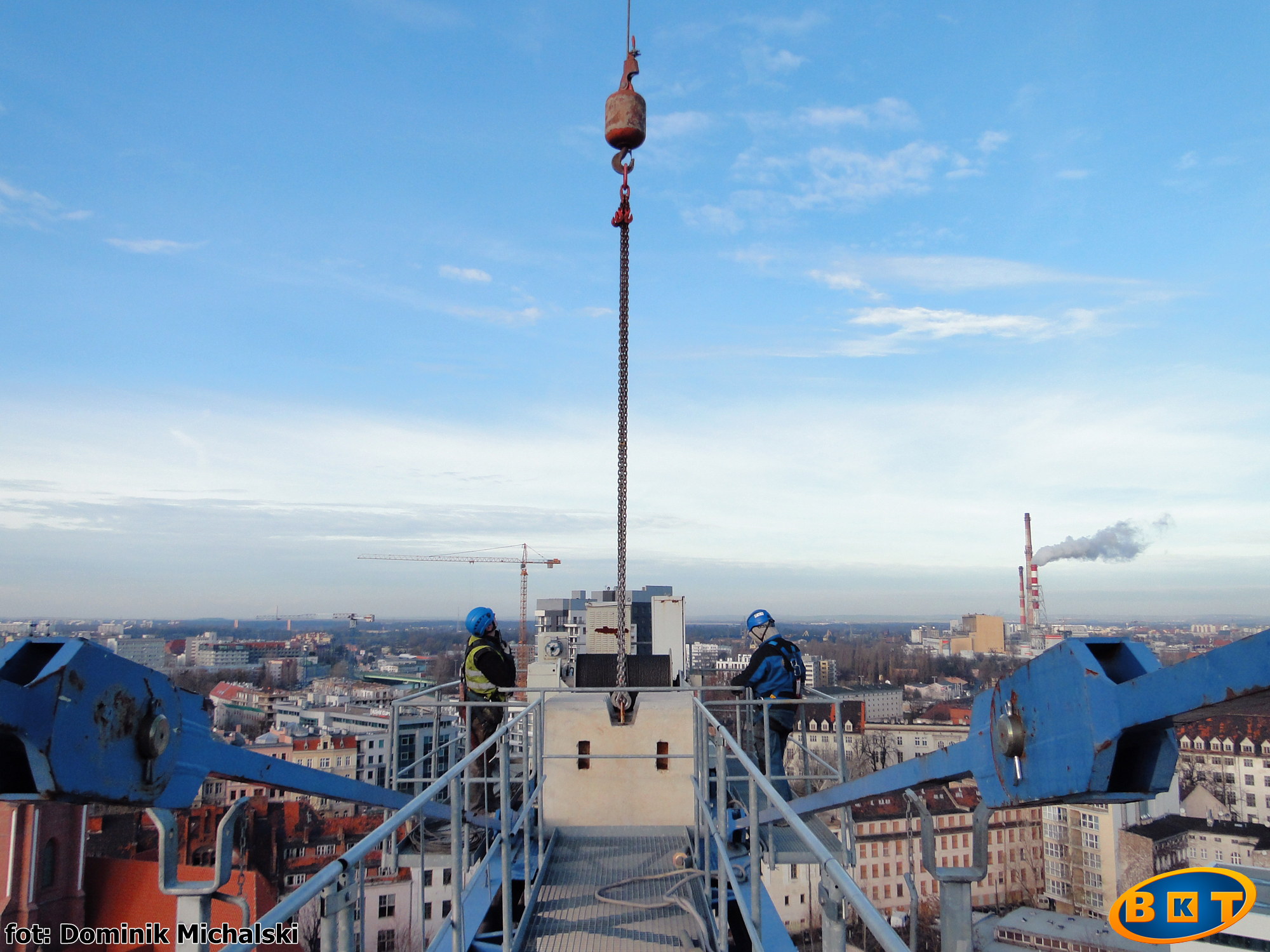 Widok z góry na dwóch pracowników w kaskach na platformie dźwigu, z widoczną panoramą miasta w tle i łańcuchem z hakiem opuszczonym w dół.