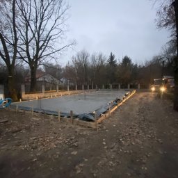 NIERUCHOMOŚCI WIPOD SPÓŁKA Z OGRANICZONĄ ODPOWIEDZIALNOŚCIĄ SPÓŁKA KOMANDYTOWA - Wylany fundament pod budowę domu, widoczne szalunki i wystające pręty zbrojeniowe, w tle drzewa bez liści i budynek mieszkalny, oświetlone reflektorami samochodu dostawczego.
