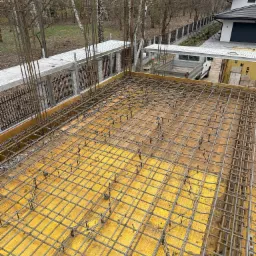 Zbrojenie płyty fundamentowej na żółtym szalunku z widocznymi pionowymi prętami i elementami dystansowymi, w tle dom jednorodzinny i drzewa.