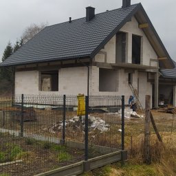 Jarosław Gruba Usługi Ogólnobudowlane - Surowy stan domu z białego bloczka z czarnym dachem z dachówki, widoczny pracownik na drabinie oraz metalowe ogrodzenie i rów melioracyjny na pierwszym planie.