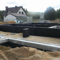 Zabezpieczone, zaizolowane fundamenty domu jednorodzinnego w trakcie budowy, z widoczną hydroizolacją i drenażem. W tle dom w stanie surowym otwartym.
