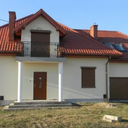 Nowoczesny dom jednorodzinny z balkonem i czerwonym dachem, brązowe rolety w oknach, garaż z dwoma bramami, jasna elewacja, słoneczny dzień.