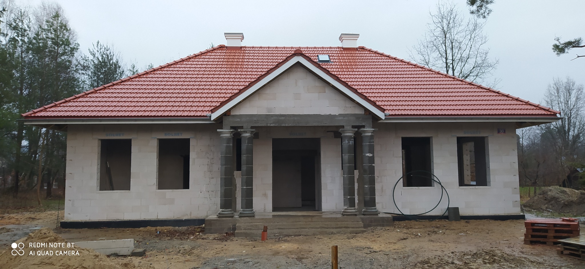 Surowy stan domu z czerwonym dachem, kolumnami przy wejściu i widocznymi pustakami Solbet, otoczonego ziemią i paletami, w pochmurny dzień.