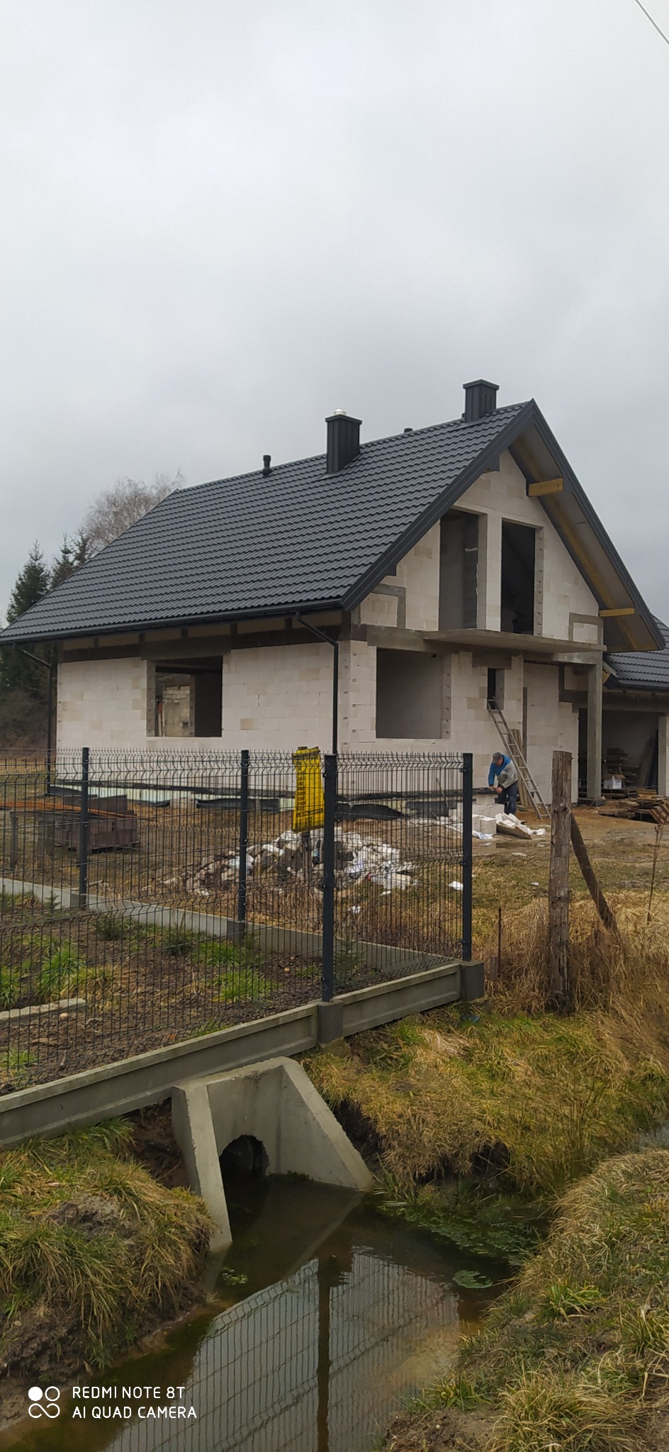 Surowy stan domu z białego bloczka z czarnym dachem z dachówki, widoczny pracownik na drabinie oraz metalowe ogrodzenie i rów melioracyjny na pierwszym planie.