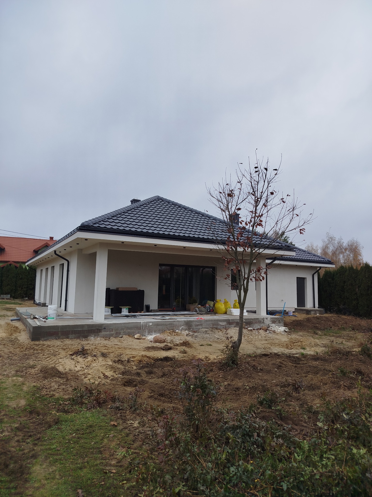 Parterowy dom z czarną dachówką w trakcie wykończenia elewacji, widoczny fundament z bloczków betonowych i teren wokół w trakcie prac ziemnych.