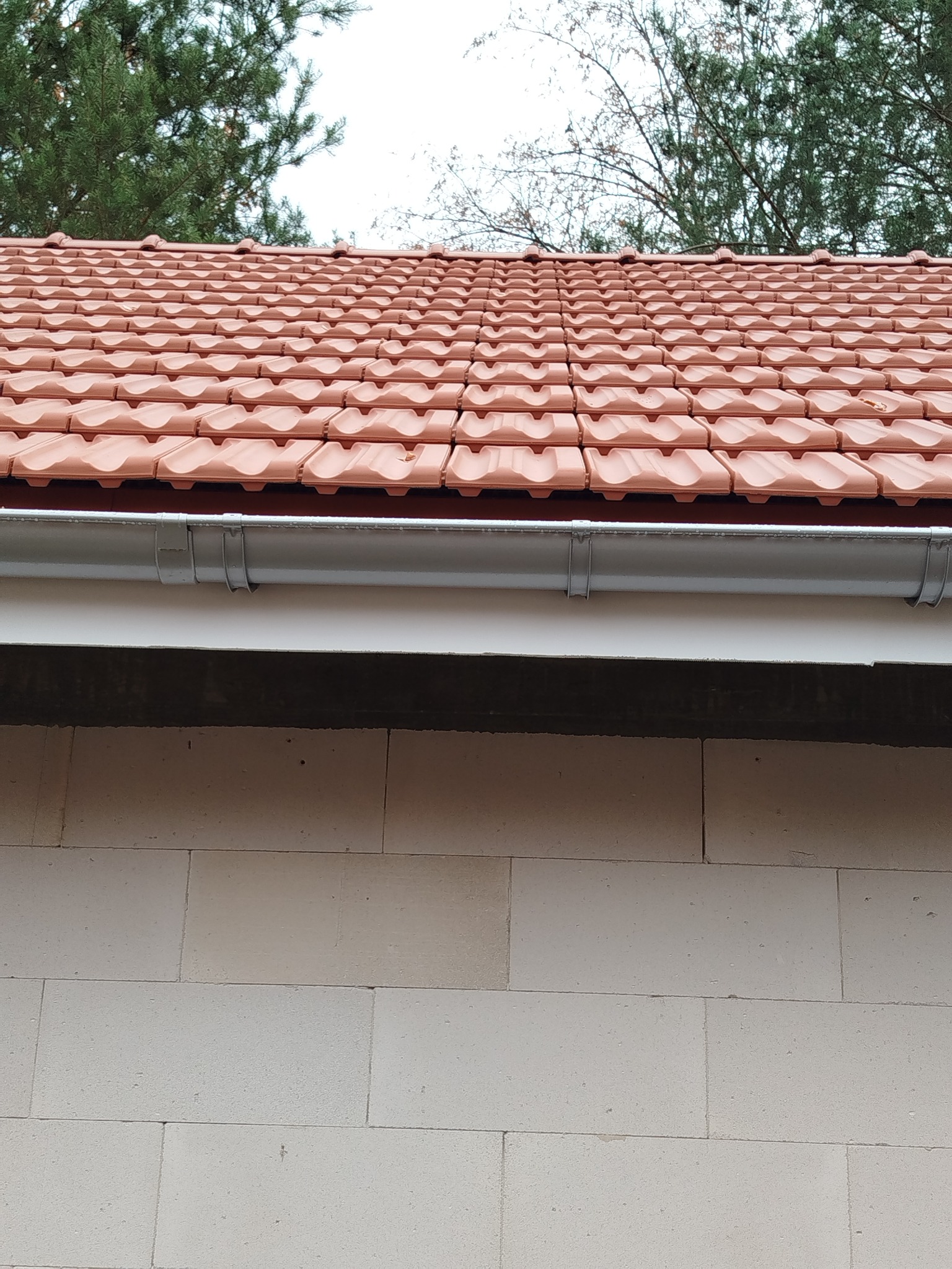 Fragment nowo wybudowanego domu z czerwonej dachówki ceramicznej, widoczny szary system rynnowy i białe bloczki silikatowe ściany, tło stanowią drzewa.