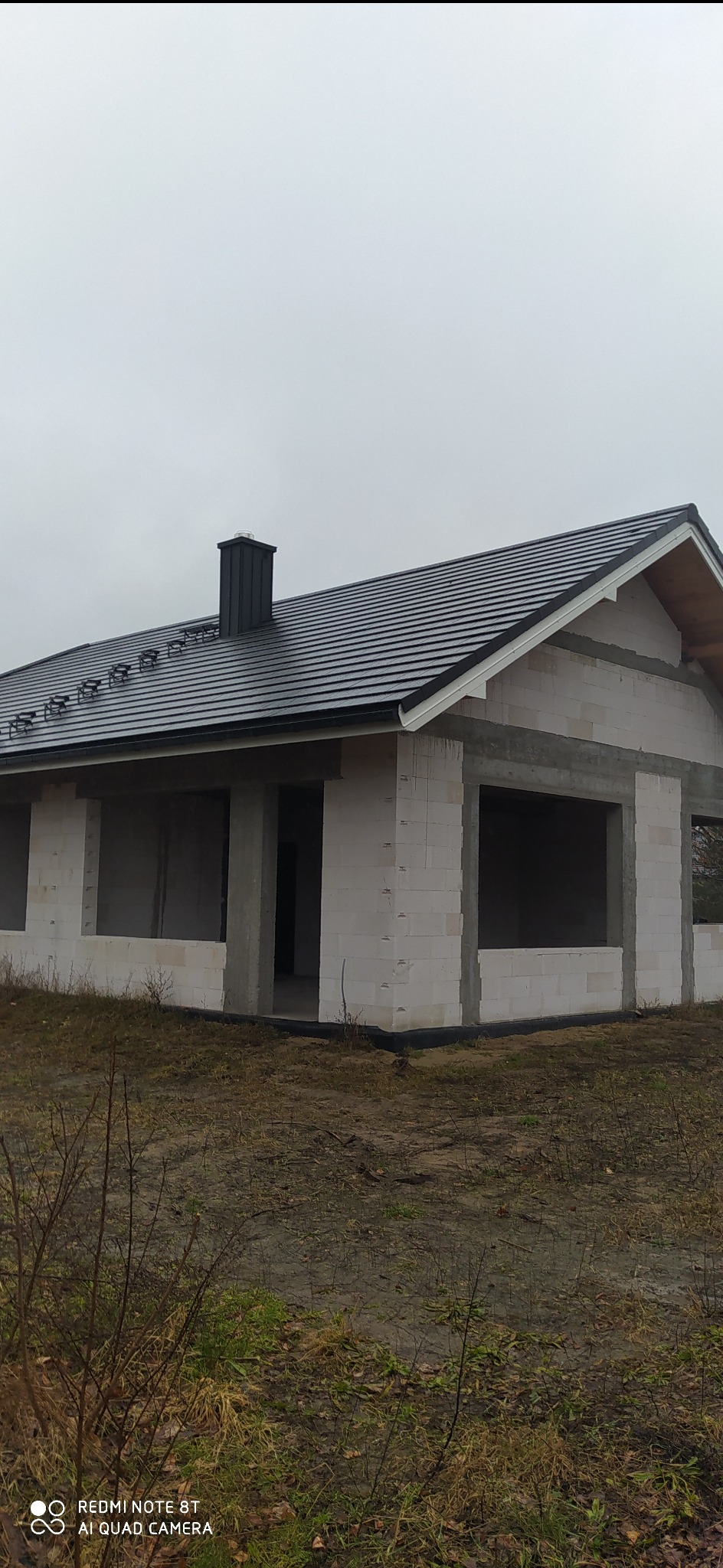 Surowy stan budynku z białych bloczków, z czarnym dachem i kominem, widoczne niezabudowane otwory okienne i drzwiowe, teren wokół niezagospodarowany, pochmurne niebo.