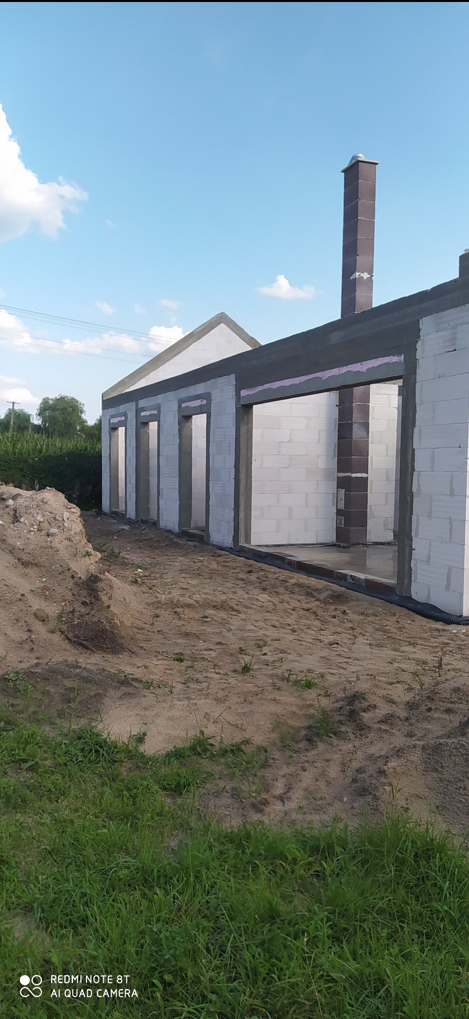 Stan surowy otwarty domu z białych bloczków, widoczne otwory okienne i drzwiowe, komin z cegły klinkierowej, grunt piaszczysty.