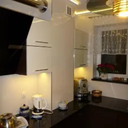 Nowoczesna kuchnia z czarnym blatem granitowym, białymi szafkami i oświetleniem LED podszafkowym, widoczne akcesoria kuchenne i kwiaty w oknie.