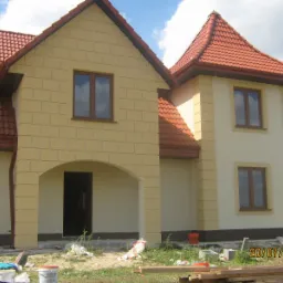 Dwukondygnacyjny dom z żółtą elewacją i czerwoną dachówką, widok z zewnątrz, teren wokół domu w trakcie prac wykończeniowych, widoczne materiały budowlane.