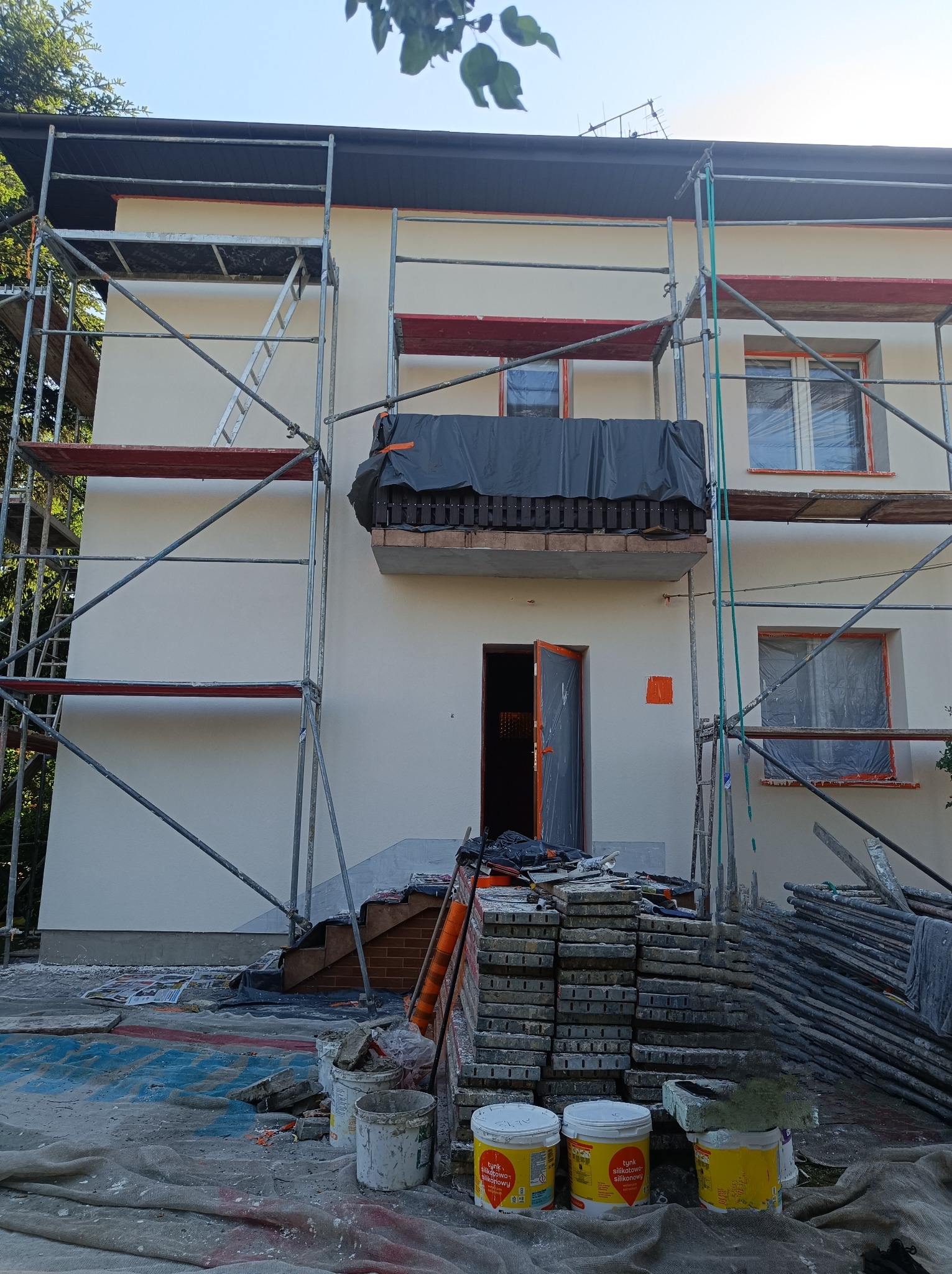 Dom w trakcie prac elewacyjnych, widoczne rusztowania, zakryty balkon, przygotowane materiały budowlane i farby, świeżo otynkowana ściana w jasnym odcieniu.