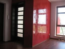 Czerwona ściana z efektem dekoracyjnym odbijającym światło, widoczna część drzwi wejściowych z przeszkleniami oraz okno z widoczną drabiną opartą o ścianę, parkiet na podłodze.