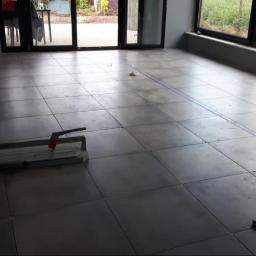 Landmen Firma ogólnobudowlana - Szare płytki podłogowe ułożone w dużym pomieszczeniu z widocznym narzędziem do cięcia płytek, niebieska linia wyznacza dalszy obszar do ułożenia płytek.