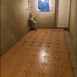 Landmen Firma ogólnobudowlana - Układanie brązowych płytek podłogowych w pomieszczeniu z szarą ścianą, widoczne krzyżyki dystansowe, okno zabezpieczone folią, drzwi po prawej stronie.