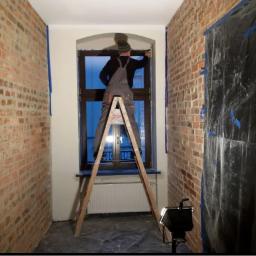 Landmen Firma ogólnobudowlana - Pracownik w kombinezonie na drabinie maluje ramę okienną w pomieszczeniu z odsłoniętą cegłą, zabezpieczoną folią i taśmą malarską.