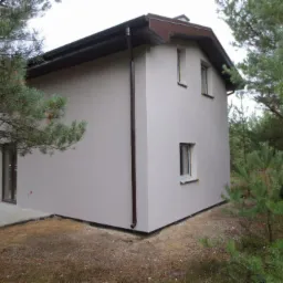 Dwukondygnacyjny dom z szarą elewacją, brązowym dachem i rynnami, widziany z niskiej perspektywy, otoczony drzewami iglastymi.