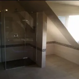 Wykończona łazienka na poddaszu ze szklaną kabiną prysznicową typu walk-in, jasnymi płytkami i oknem w skośnej ścianie.