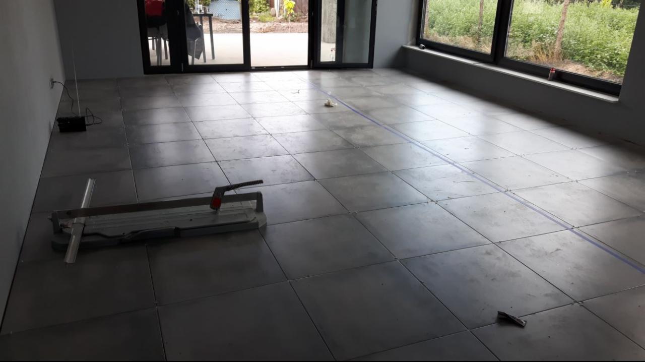 Szare płytki podłogowe ułożone w dużym pomieszczeniu z widocznym narzędziem do cięcia płytek, niebieska linia wyznacza dalszy obszar do ułożenia płytek.