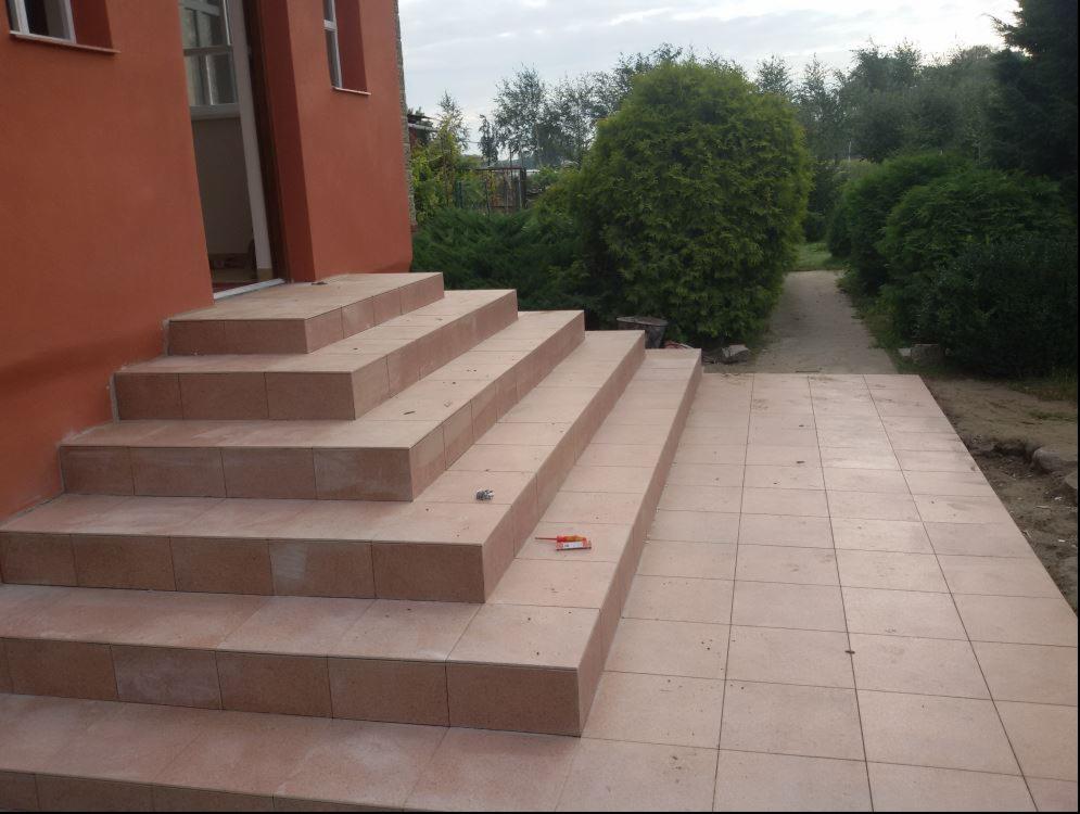 Wykończone kafelkami schody wejściowe do domu z pomarańczową elewacją, widoczny fragment terakoty przed schodami i ścieżka w ogrodzie w tle, na jednym ze stopni leży śrubokręt.