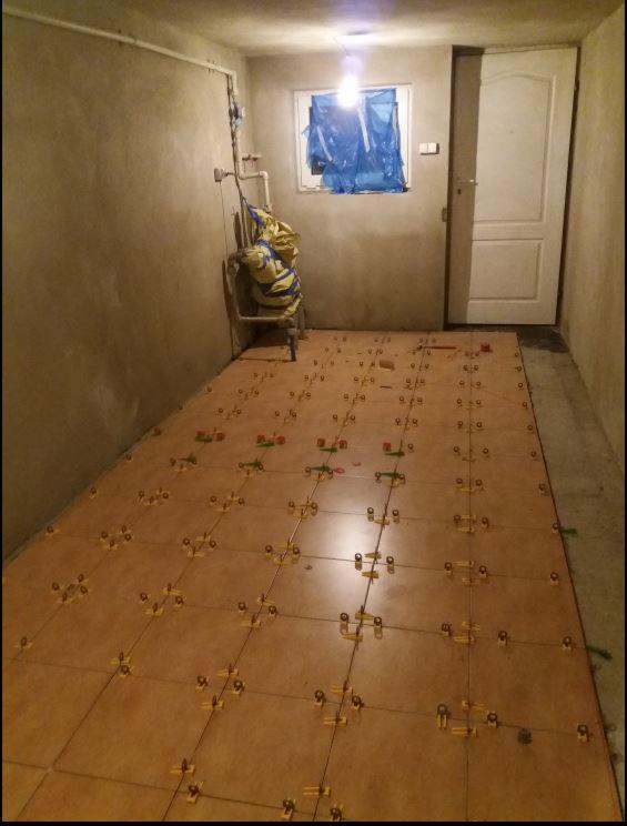 Układanie brązowych płytek podłogowych w pomieszczeniu z szarą ścianą, widoczne krzyżyki dystansowe, okno zabezpieczone folią, drzwi po prawej stronie.