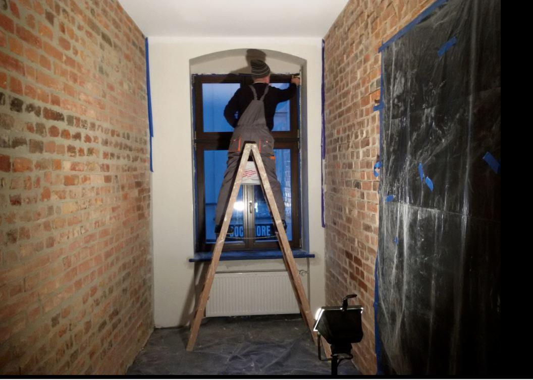 Pracownik w kombinezonie na drabinie maluje ramę okienną w pomieszczeniu z odsłoniętą cegłą, zabezpieczoną folią i taśmą malarską.