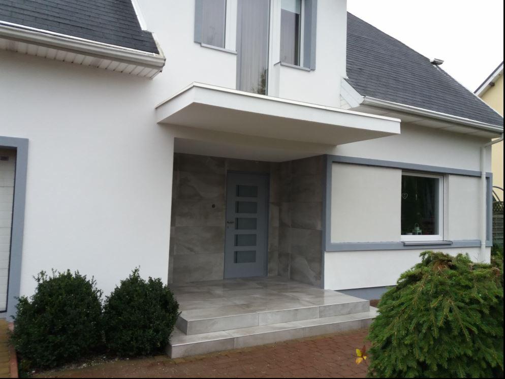 Wejście do domu jednorodzinnego z szarą stolarką okienną i drzwiową, wykończone płytami imitującymi kamień, zadaszone płaskim daszkiem, z dwoma stopniami wejściowymi i niską zielenią.