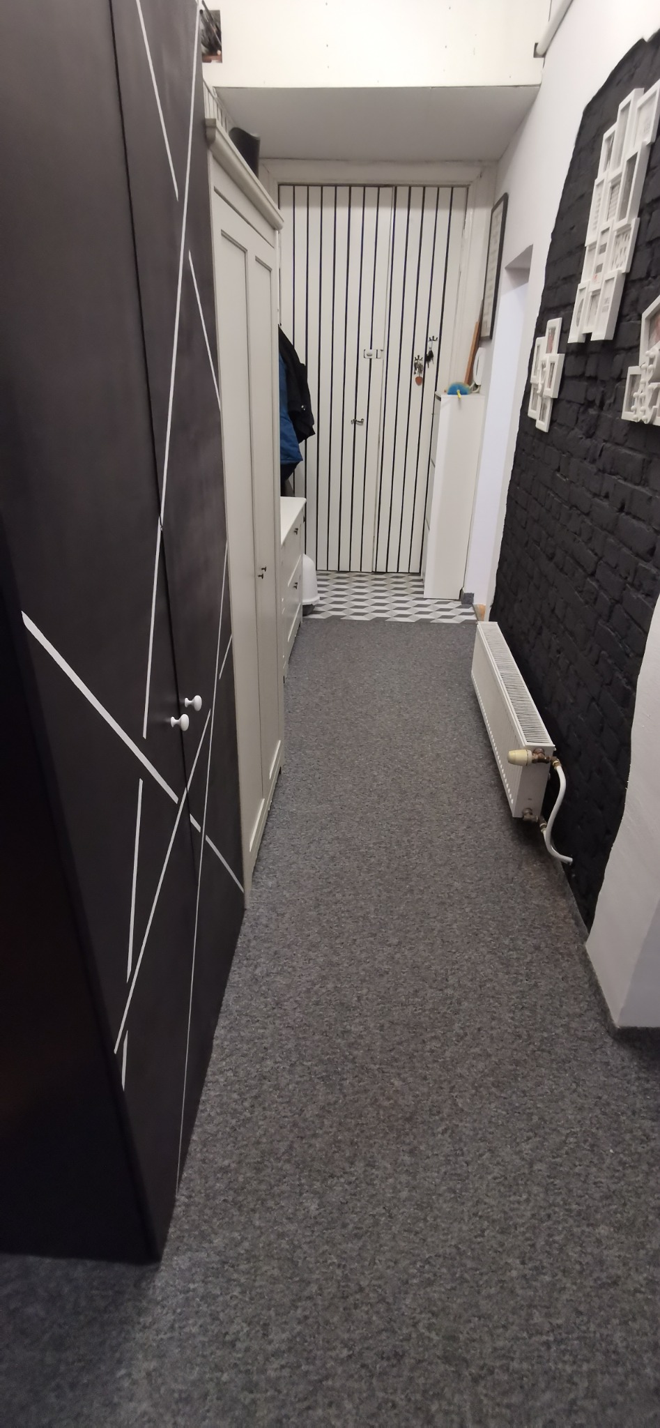 Wnętrze korytarza z ciemną szafą z białymi geometrycznymi wzorami, białą komodą, grzejnikiem i czarną ścianą z dekoracyjnymi ramkami.