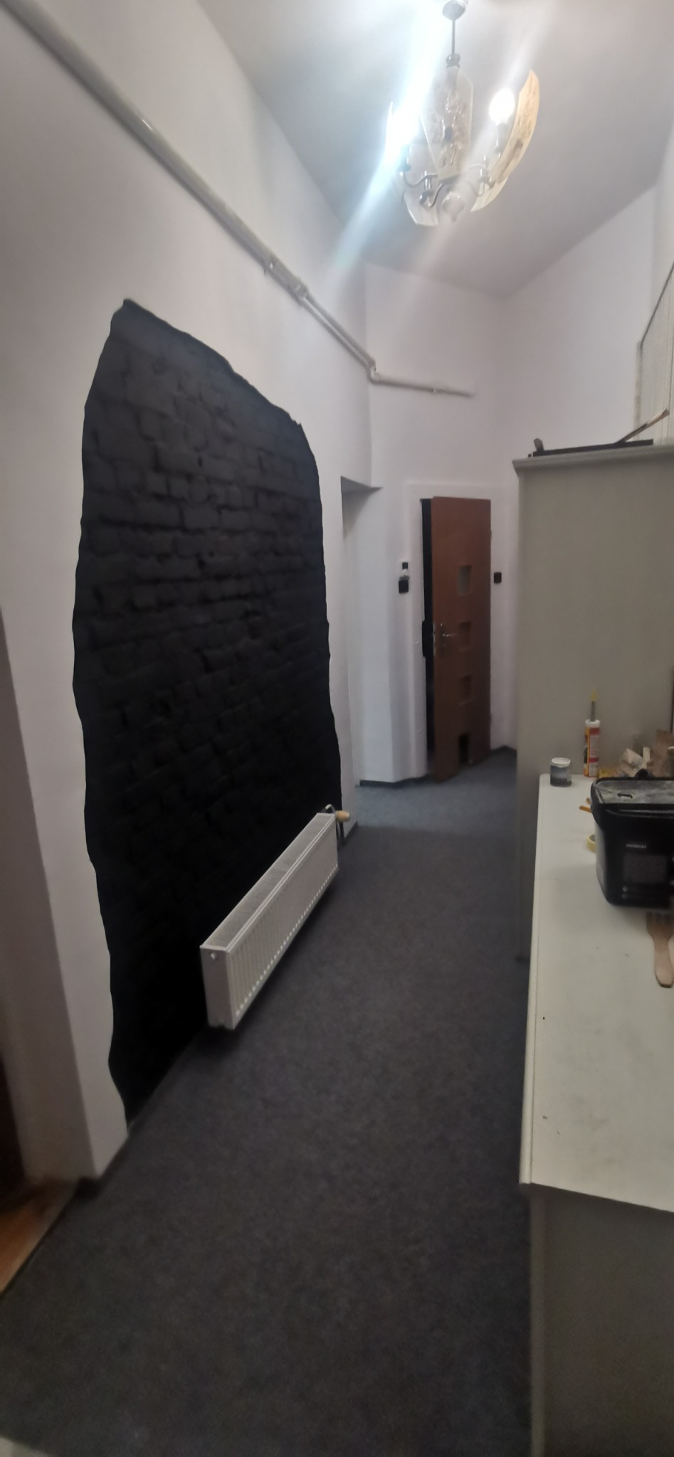 Ciemna, nieregularna plama imitująca cegłę na białej ścianie w korytarzu, obok grzejnik, widok na drzwi i fragment szafki z akcesoriami.