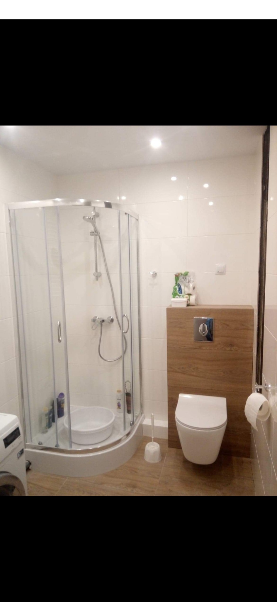 Nowoczesna łazienka z kabiną prysznicową narożną, podwieszaną toaletą z drewnianą obudową spłuczki i pralką w rogu, wykończona jasnymi płytkami.