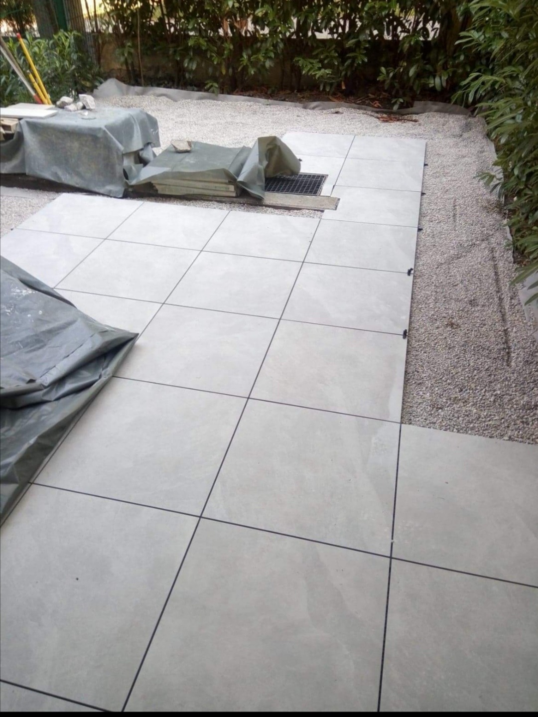 Układanie jasnych, kwadratowych płytek na zewnątrz, z widocznym drenażem i podsypką z drobnego żwiru, częściowo przykryte folią ochronną.