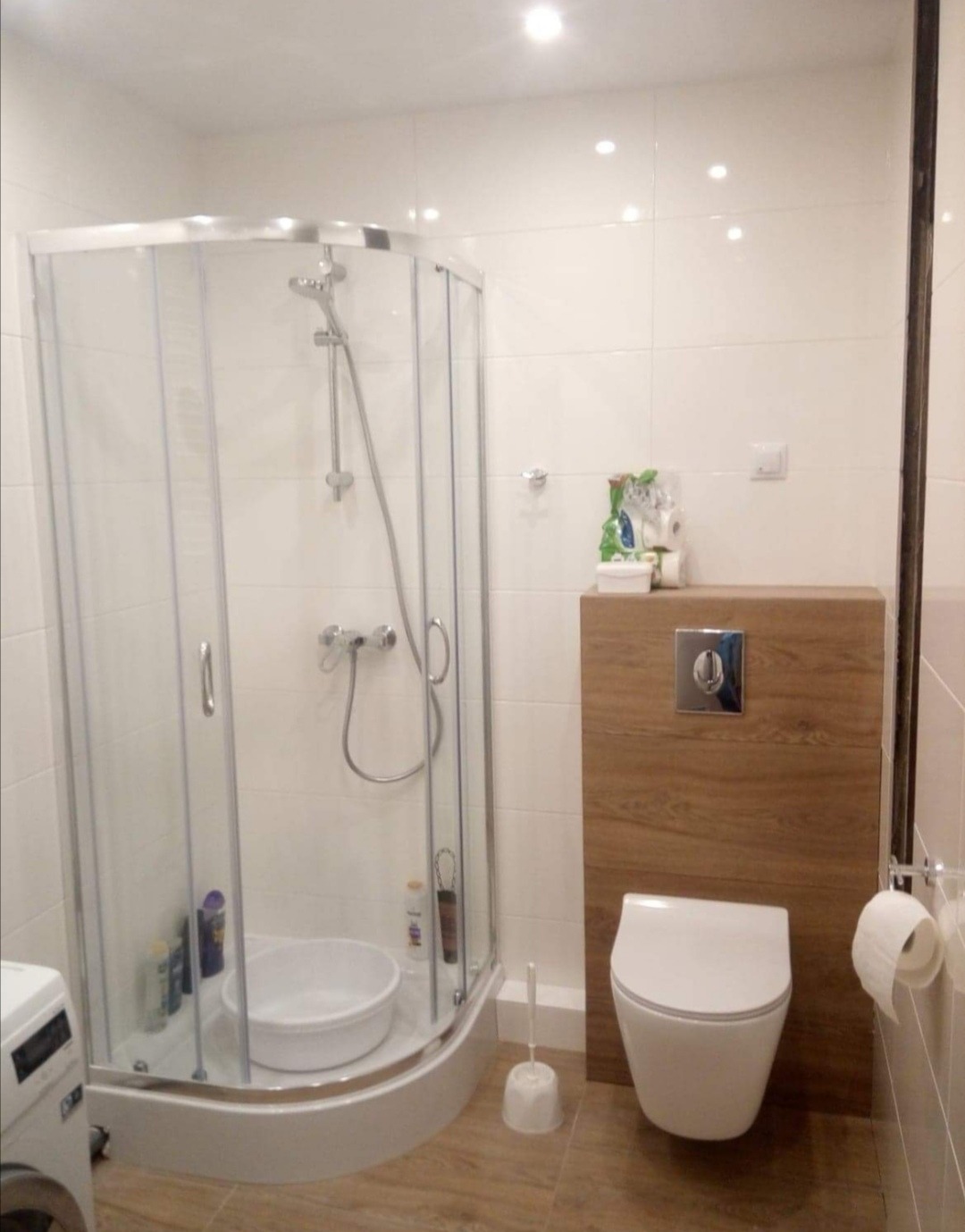 Nowoczesna łazienka z narożną kabiną prysznicową, podwieszaną toaletą i drewnopodobną zabudową, widoczny fragment pralki z lewej strony.