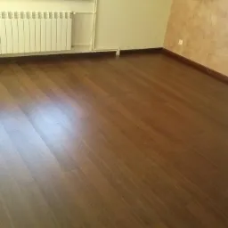 Ułożone ciemnobrązowe panele podłogowe w pustym pokoju z grzejnikiem pod oknem i beżową ścianą z dekoracyjną fakturą.