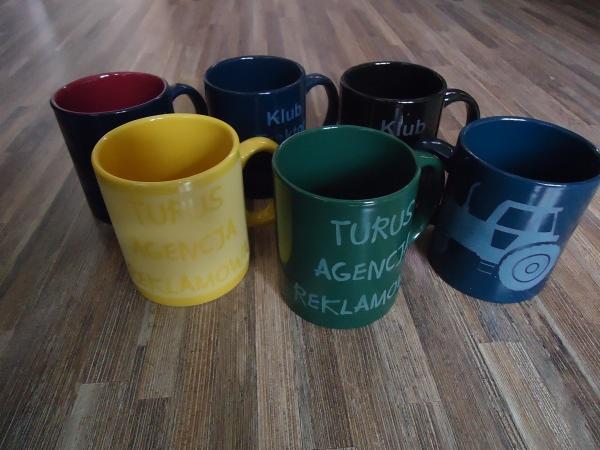 Sześć kolorowych kubków z napisami i grafikami, ustawionych na drewnopodobnej powierzchni. Kubki w kolorach czerwonym, granatowym, czarnym, żółtym, zielonym i niebieskim. Na kubkach widoczne napisy...