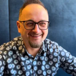 Uśmiechnięty mężczyzna w okularach i koszuli w kwiaty siedzi na tle niebieskiej tapicerki.