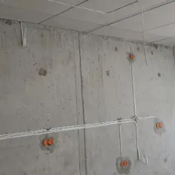 Surowy betonowy pokój w trakcie remontu, z widocznymi białymi przewodami elektrycznymi w peszlach, wystającymi ze ścian i sufitu, oraz pomarańczowymi puszkami instalacyjnymi. Metalowa rura...