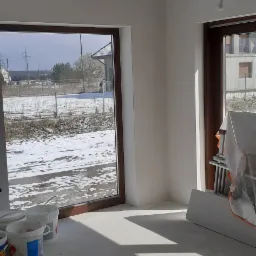Wnętrze po szpachlowaniu i gładziowaniu ścian, widoczne wiaderka po materiałach, okna z widokiem na zimowy krajobraz.