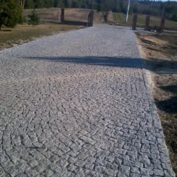 Szara, granitowa ścieżka z elementami ułożonymi w okrągłe wzory, prowadząca przez trawnik w kierunku lasu. Widoczne słupki z cegły po bokach ścieżki.