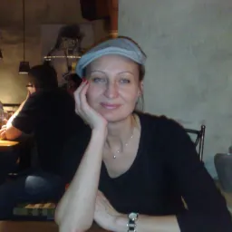 Kobieta w szarej czapce siedzi przy stole w restauracji, wspierając głowę na dłoni i uśmiechając się.