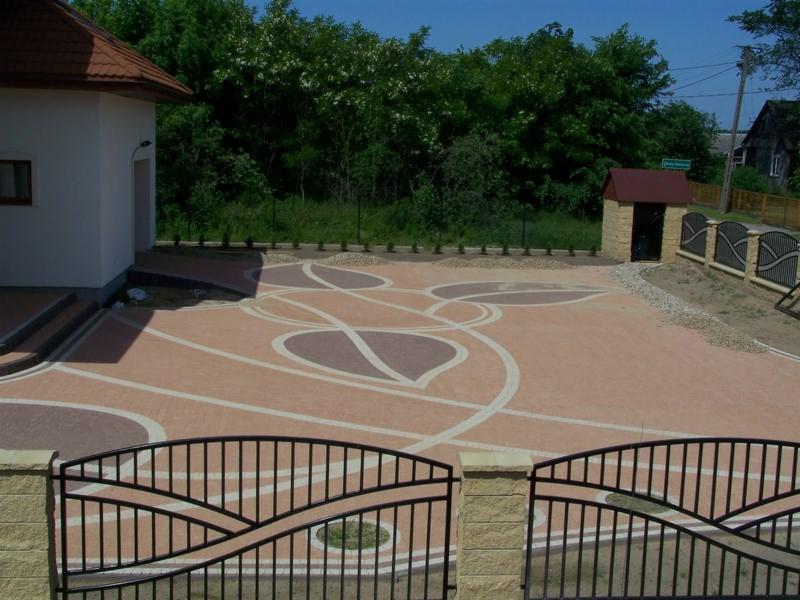 Ułożona nawierzchnia z kostki brukowej w odcieniach beżu i brązu z dekoracyjnym wzorem przypominającym liście, widok zza czarnej metalowej bramy, w tle dom i mała murowana budka.