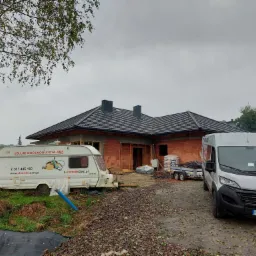 Dom w trakcie budowy z czerwonej cegły, widoczny szary dach, przyczepa kempingowa z reklamą usług budowlanych oraz szary samochód dostawczy na tle pochmurnego nieba.