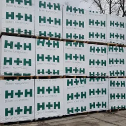 Palety z białymi opakowaniami betonu komórkowego H+H, składowane na placu budowlanym, widoczne drzewa w tle.