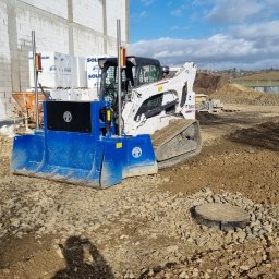 BEAR Polska - Biała miniładowarka Bobcat z niebieskim osprzętem do równania terenu, obok walec drogowy ubijający ziemię, w tle budynek w trakcie budowy i pagórkowaty krajobraz.