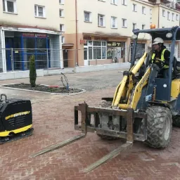 Żółta mini-ładowarka Mustang z widłami i zagęszczarka Bomag na nowo ułożonej, czerwonej kostce brukowej w centrum miasta, widoczny pracownik w kasku i kamizelce odblaskowej.